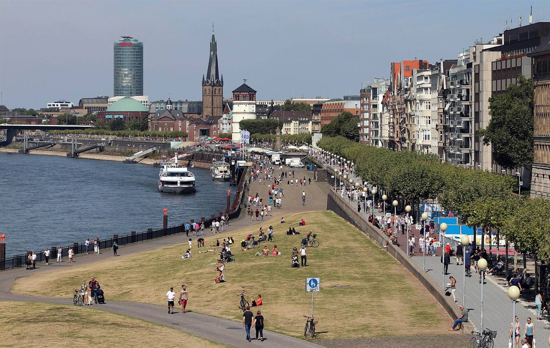 Sommertag an der Düsseldorfer Rheinuferpromenade: Menschen sitzen auf der Wiese, spazieren und fahren Rad, dahinter Altstadt und Schlossturm
