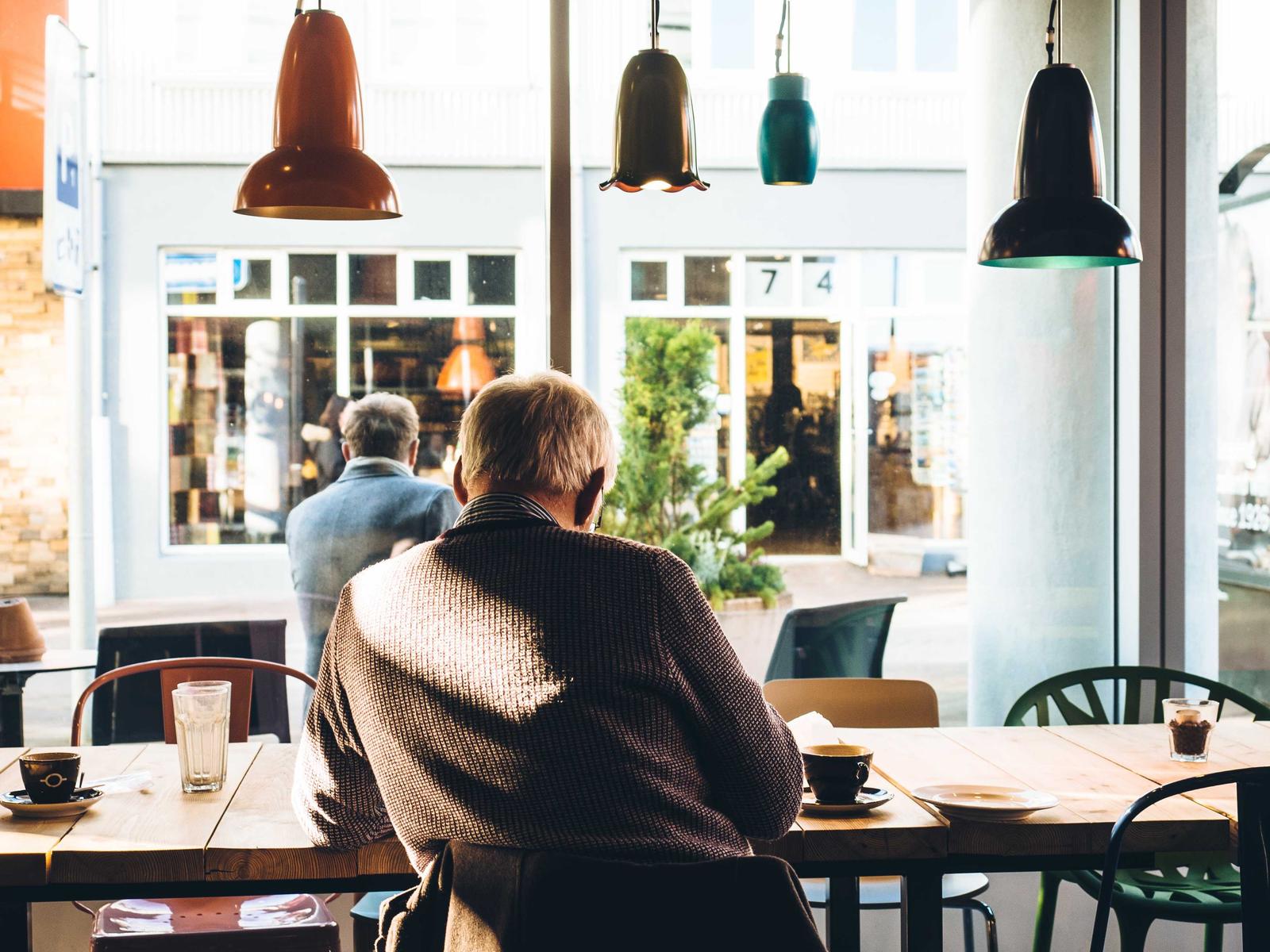 Älterer Mann sitzt mit einem Kaffee an einem hellen Fenster im Café
